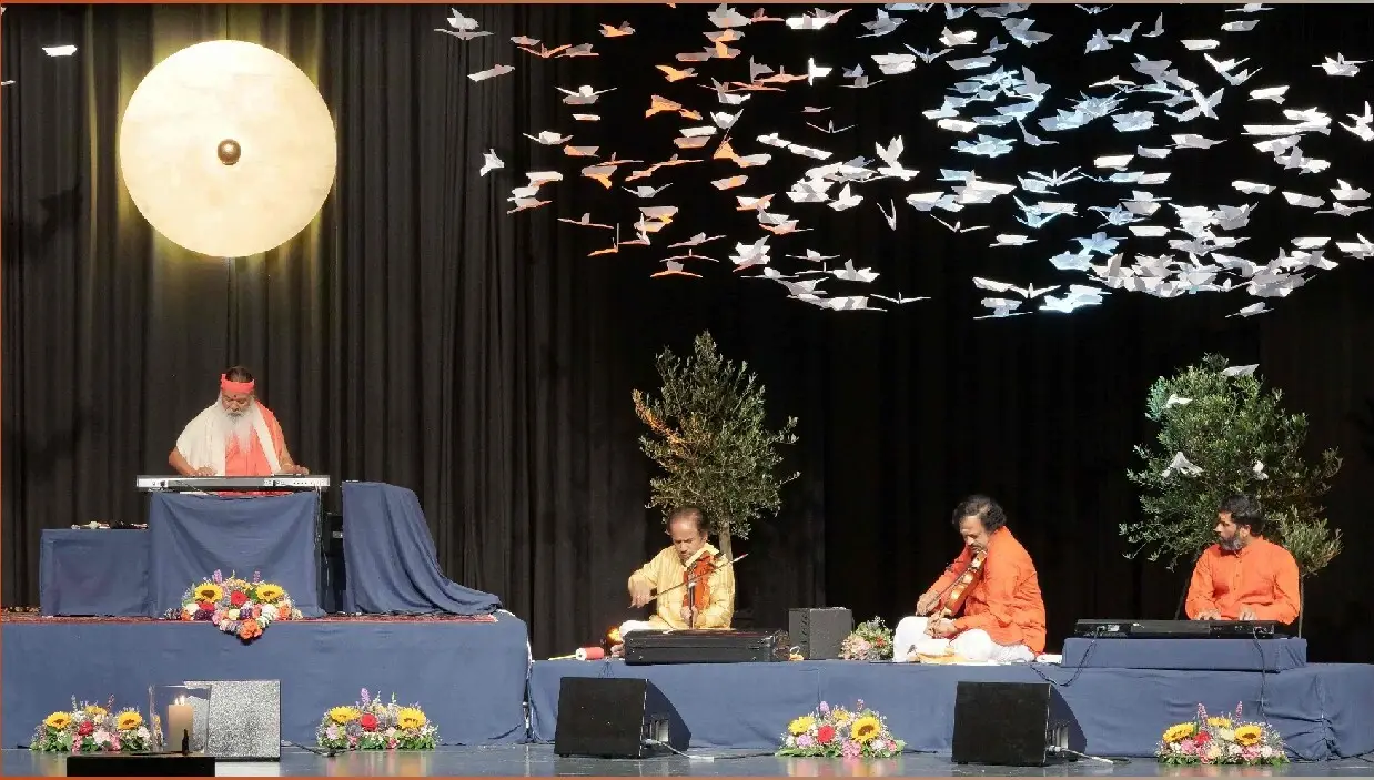 Sri Swamiji and ensemble on stage: synthesiser, violin, mridangam and tabla, performing under hundreds of suspended paper birds.